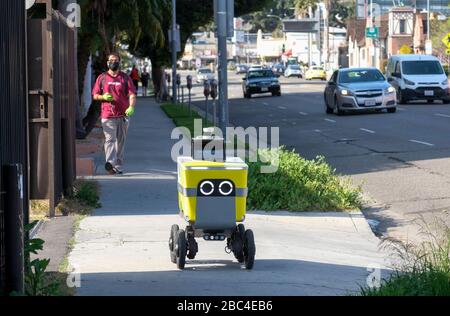Los Angeles, Kalifornien, USA. April 2020. Ein Mann, der eine Gesichtsmaske trägt, läuft hinter einem Lebensmittel-Lieferroboter, als er sich am 2. April 2020 in Los Angeles auf den Weg macht. Der Bürgermeister von Los Angeles, Eric Garcetti, rät der Öffentlichkeit, beim Aussteigen eine Schutzhülle zu tragen - auch einen Schal oder ein anderes Kleidungsstück - um die Ausbreitung des Coronavirus zu verringern. Kredit: Ringo Chiu/ZUMA Wire/Alamy Live News Stockfoto