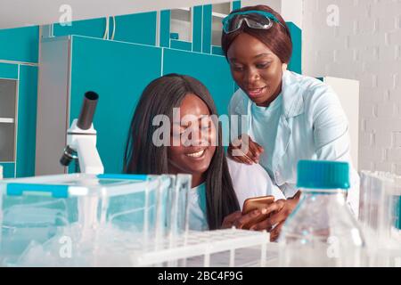 Zwei junge lächelnde afrikanische Wissenschaftlerinnen, Medizinstudenten mit Handy. Frauen lächeln und lachen über etwas auf dem Smartphone. Wissenschaftliche Chemie Stockfoto