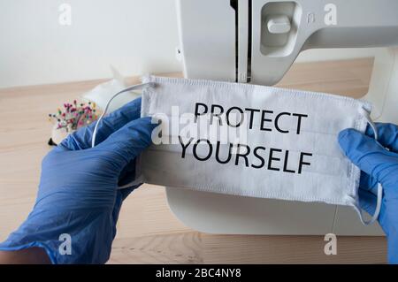 Schützen Sie sich vor Coronavirus, und nähen Sie zu Hause eine medizinische Gesichtsmaske. Hände in blauen Handschuhen, die Maske mit Schild halten. Stockfoto