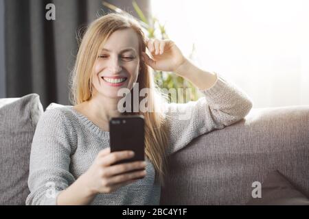 Lächelnde, reife Frau im häuslichen Outfit auf der Couch sitzen und Videokonferenzen auf dem Smartphone abhalten. Schöne Dame mit blonden Haaren, die Freizeit verbringt ti Stockfoto