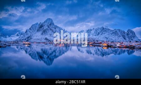 Die Abendlichter des Dorfes Reine, mit schneebedeckter Bergkette im Rücken, und seine Reflexion im ruhigen Wasser des Fjords. Stockfoto