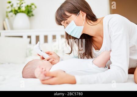 Mutter mit Gesichtsmaske und Baby mit Fiebermaß mit Ohrthermometer Stockfoto