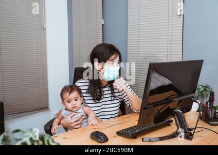 Coronavirus. Geschäftsfrau, die von zu Hause aus mit Schutzmaske arbeitet. Geschäftsfrau in Quarantäne arbeitet während der Babysitting Stockfoto