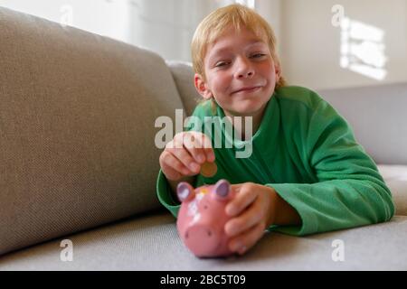 Fröhlicher junger gutaussehender Junge, der zu Hause eine Münze auf der Schweinebank einsteckte Stockfoto