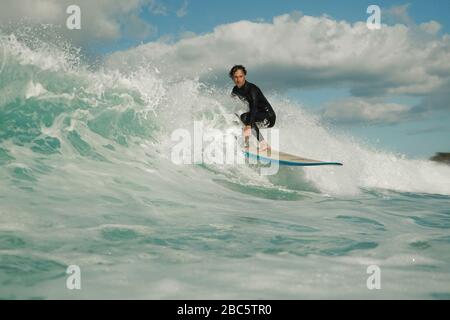 Ein junger männlicher Surfer in einem schwarzen Neoprenanzug reitet auf einer Bruchwelle mit weißem Wasser ein Longbord-Surfbrett. Sommertag mit blauem bewölktem Himmel. Stockfoto