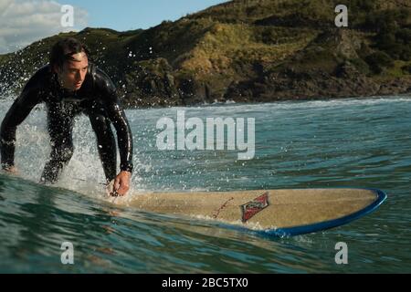 Ein junger männlicher Surfer in einem schwarzen Neoprenanzug reitet auf einer Bruchwelle mit weißem Wasser ein Longbord-Surfbrett. Sommertag mit blauem bewölktem Himmel. Stockfoto