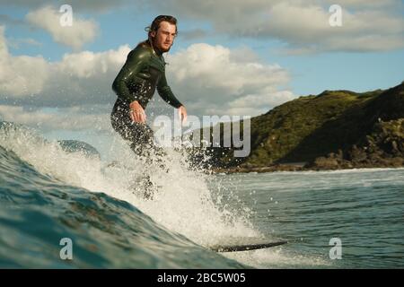 Ein junger männlicher Surfer in einem schwarzen Neoprenanzug reitet auf einer Bruchwelle mit weißem Wasser ein Longbord-Surfbrett. Sommertag mit blauem bewölktem Himmel. Stockfoto