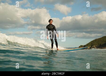 Ein junger männlicher Surfer in einem schwarzen Neoprenanzug reitet auf einer Bruchwelle mit weißem Wasser ein Longbord-Surfbrett. Sommertag mit blauem bewölktem Himmel. Stockfoto