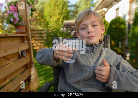 Junger gutaussehender Junge mit einem Glas Wasser, das im Hinterhof Daumen nach oben gibt Stockfoto