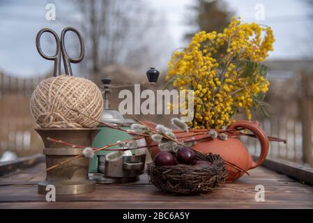 Auf der Holztischplatte befindet sich ein Gelege mit bemalten Wachteleiern, Weidenästen und verschiedenen Haushaltsgeräten. Stockfoto