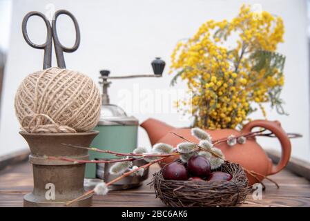 Auf der Holztischplatte befindet sich ein Gelege mit bemalten Wachteleiern, Weidenästen und verschiedenen Haushaltsgeräten. Stockfoto