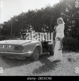 1970er Jahre, historisch, draußen auf einem sonnenbeleuchteten Feld, eine attraktive blonde Frau in einer weißen Oberteil- und glockenuntergebrachten Hose, neben ihrem offenen Sportwagen, einem Triumph Spitfire IV, mit Drahträdern, England, Großbritannien stehend. Der 1962 erstmals vorgestellte Spitfire, der auf einem Entwurf von Giovanni Michelotti basierte, war ein kleiner britischer Zweisitzer-Sportwagen und wurde bis 1980 hergestellt. Das hier abgebildete Mark IV-Modell wurde in den Jahren 1970 bis 1974 bei Standard-Triumph Works (im Besitz von Leyland Motors) in Canley, Coventry hergestellt und neben anderen neuen Merkmalen aus früheren Modellen war ein neu gestaltetes Heckende Stockfoto