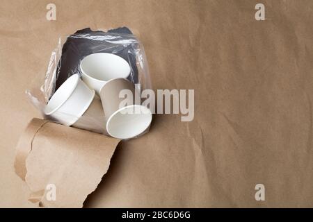 Umweltfreundliche, kompostierbare, recycelbare Papierbecher in Plastiktüten durch das Loch im braunen Papierhintergrund. Bodenverschmutzungskonzept. Erstickt Stockfoto