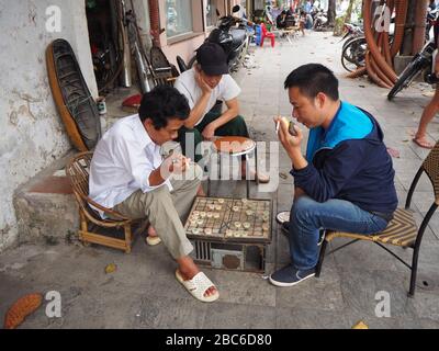 3 lokale Männer, die Schach im vietnamesischen Stil (Checkers) auf dem Straßenbelag in einer Straße in Hanoi, Vietnam spielen Stockfoto