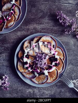 Violette Pfannkuchen mit Blaubeeren und Creme, die im Frühling mit lilakalen Blumen im dunklen Hintergrund dekoriert sind Stockfoto