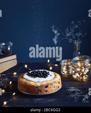 Hausgemachter Kuchen mit schwarzem Currant und Mascarpone auf dem Holztisch in der Nähe alter Bücher und Glasgefäße mit hellen Sternen auf dunklem Hintergrund im Sommer Stockfoto