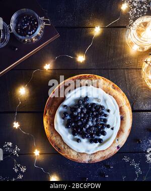 Hausgemachter Kuchen mit schwarzem Currant und Mascarpone auf dem Holztisch in der Nähe von Buch- und Glasgefäßen mit hellen Sternen auf dunklem Hintergrund im Sommer Top VI Stockfoto