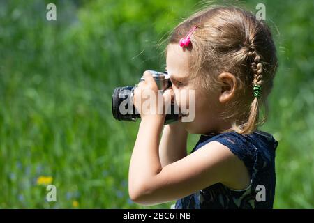 Ein kleines Mädchen macht sich im Sommer im Park mit einer Retro-Filmkamera ein Bild. Kleines hübsches Kindermädchen in leichtem Kleid mit Retro-Vintage Stockfoto