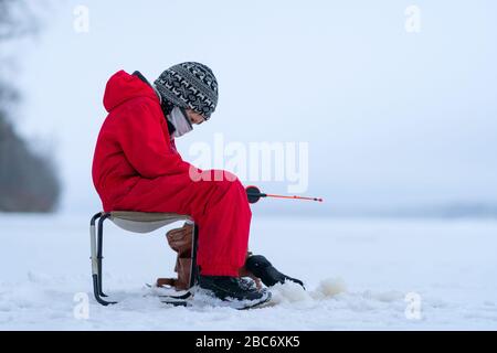 Ein Junge in roten Overalls beim Winterangeln auf dem See. Sitzt in einem Sessel in der Nähe des Lochs. In den Händen einer Angelrute. Schaut vorsichtig auf die Angelrute. Der Horizont verschmilzt mit dem Himmel. Stockfoto