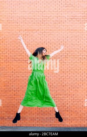 Frau in einem grünen Kleid, die vor einer Ziegelwand glücklich auf und ab springt. Stockfoto