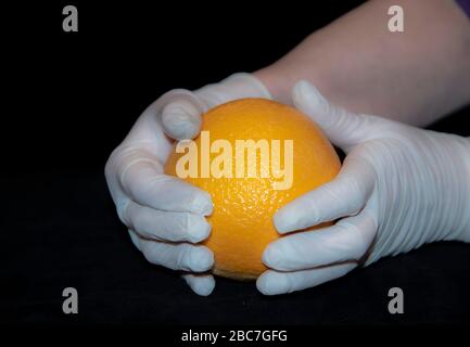 Die Hände einer älteren Frau in weißen Gummihandschuhen halten eine große orange Orange. Stockfoto