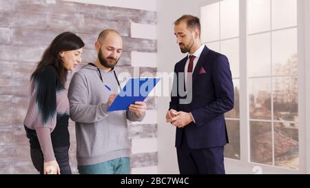 Junge Paare werden Hausbesitzer, nachdem sie Dokumente mit Immobilienmakler in Geschäftsanzug unterschrieben haben. Agent gibt Schlüssel zum Paar. Stockfoto