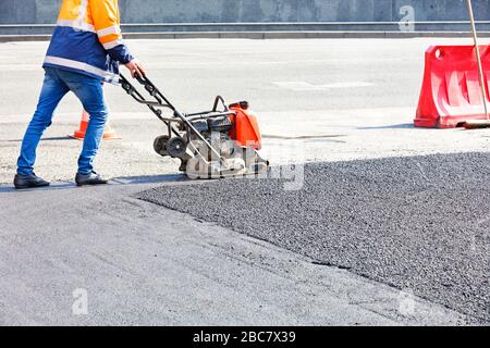 Der Straßendienst des Fahrers nutzt den vibrierenden Plattenverdichter, der Asphalt am Straßenreparaturstandort verdichtet. Stockfoto