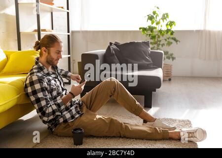 Foto des jungen gutaussehenden Mannes, der ein Plaid Hemd trägt, das Handy hält und auf dem Boden in der Wohnung sitzt Stockfoto