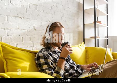 Foto eines jungen, gutaussehenden Mannes, der Kopfhörer mit einem Laptop-Computer trägt, während er auf dem Sofa in der Wohnung sitzt Stockfoto