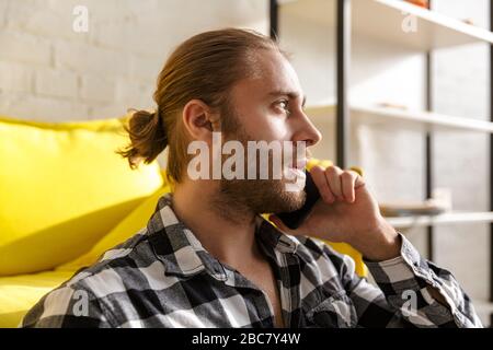 Foto des jungen gutaussehenden Mannes, der ein Plaid Hemd trägt, das auf dem Handy spricht und auf dem Boden in der Wohnung sitzt Stockfoto