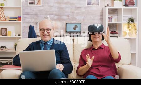 Ältere Frauen sitzen auf dem Sofa und tragen eine virtuelle Realitätsbrille. Der alte Mann sitzt auf dem Sofa mit einem Laptop neben seiner Frau Stockfoto