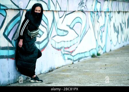 Mädchen in einer lila Maske und in einer schwarzen langen Jacke schaut auf die Kamera stehen neben einer gemalten Wand, Graffiti. Quarantäne. Covid-19 Stockfoto