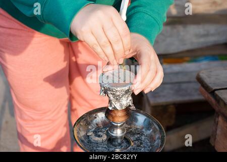 Weibliche Hand mit Hookah auf verschwommenem Hintergrund. Tasse für Hookah mit Folie gestreckt, Hände vorbereiten Hookah, Löcher in Folie machen Stockfoto