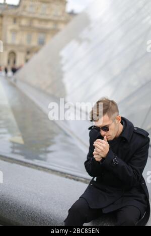 Junger Mann, der schwarze Jacke trägt, sitzt auf der Louvre Pyramide und raucht. Stockfoto