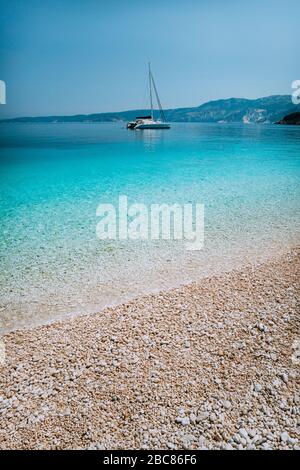 Kieselstrand mit reinem, klarem, azurblauem Wasser und Segelkatamaran-Yacht vor Anker am Horizont. Stockfoto