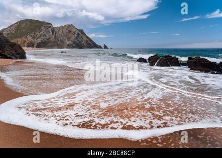 Praia da Adraga am Atlantik, Portugal. Schäumende Welle am sandigen Strand mit malerischen Landschaft Hintergrund. Stockfoto