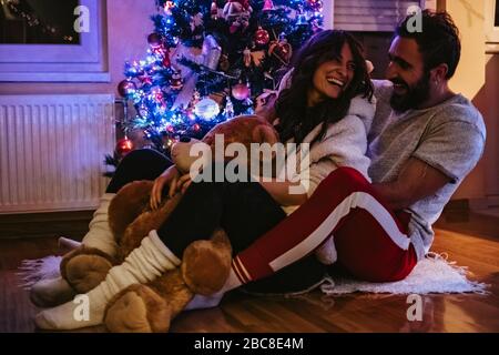 Ein Paar lacht und umarmt vor dem Weihnachtsbaum. Mädchen hält Teddybär Stockfoto