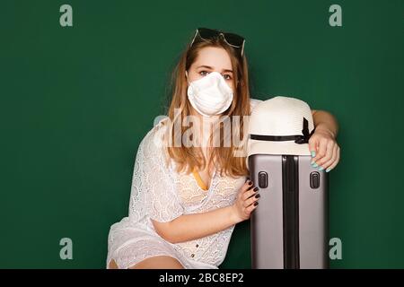 Schönes Mädchen in einem weißen Sommerkleid mit Koffer. Medizinische Maske im Gesicht. Geschlossene Ränder während des Coronavirus. Tourist auf grünem Hintergrund Stockfoto