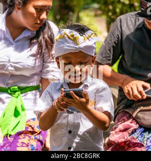 Quadratischer Blick auf einen kleinen Jungen im traditionellen Kleid mit einem Telefon in Bali, Indonesien. Stockfoto