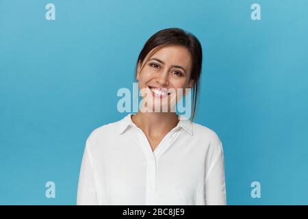 Lächelnd positive weibliche mit attraktiven Look, tragen weiße T-Shirt, posieren gegen blaue leere Wand Stockfoto