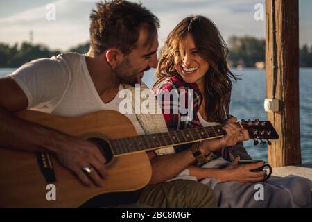 Freundin lacht, während Freund Gitarre am Fluss spielen. Wunderschöner Sonnenuntergang Stockfoto