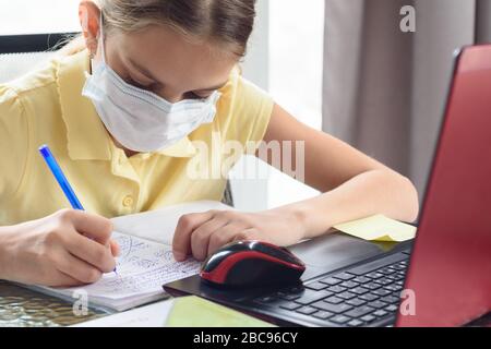 Porträt eines Mädchens in einer medizinischen Maske, das mit einem Computer am Tisch sitzende Lektionen lernt Stockfoto