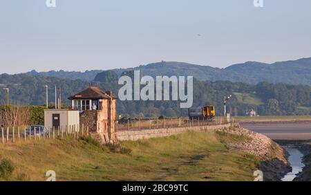 Sprinterzug der Northern Rail Klasse 156, der mit einem leeren Bestandszug am frühen Morgen an der Eisenbahnlinie der Cumbrian Coast vorbei an der Arnside Signalbox fährt Stockfoto