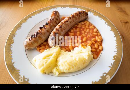 Zwei große Bratwürste mit gebackenen Bohnen und Kartoffelpüree auf einem weißen Teller mit goldenem Farbbesatz auf einem Holztisch, der von Tageslicht beleuchtet wird. Stockfoto