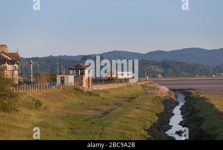 Sprinterzug der Northern Rail Klasse 156, der mit einem leeren Bestandszug am frühen Morgen an der Eisenbahnlinie der Cumbrian Coast vorbei an der Arnside Signalbox fährt Stockfoto