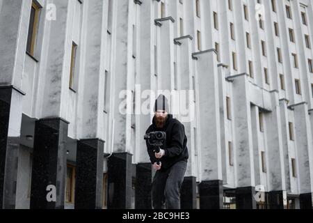 Bärtiger professioneller Videofilmer mit schwarzem Hoodie, der die professionelle Kamera auf einem 3-Achsen-Gimbal-Stabilisator hält. Filmemacher, die ein großartiges Video mit einem machen Stockfoto