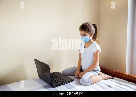 Bleiben Sie zu Hause Quarantäne Coronavirus Pandemie Prävention. Kind in medizinische Maske sitzt auf dem Bett und beim Laptop. Online-Lernen. Entfernung Stockfoto