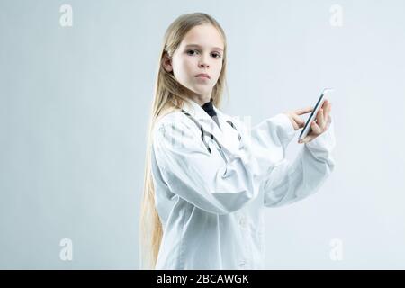 Das ernste junge Mädchen, das sich als Ärztin oder Krankenschwester verkleidet hat und ein Handy in den Händen hält, während sie sich an der auf Weiß isolierten Kamera anstarren lässt Stockfoto