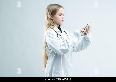 Junges Mädchen, das sich in einen medizinischen Labormantel mit Stethoskop eingekleidet hat und vorgibt, ein Arzt zu sein, der mit ihrem Handy auf weiß isoliert steht Stockfoto