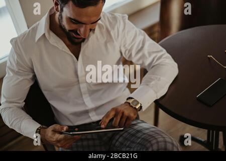 Elegant lächelnder bärtiger Mann, der auf die Tablette schaut, während er in einem Café sitzt Stockfoto